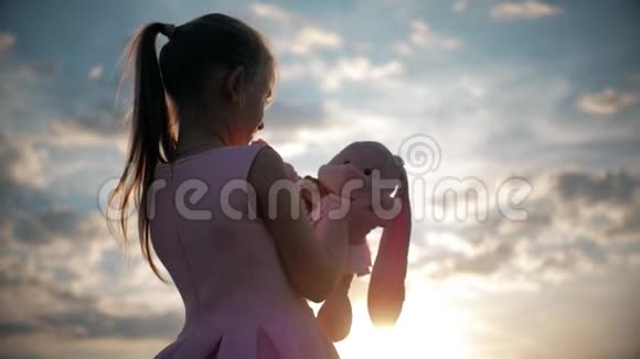 小女孩穿着粉红色的节日礼服在日落时拿着大毛绒兔子玩具一个带玩具的孩子的剪影纺织视频的预览图