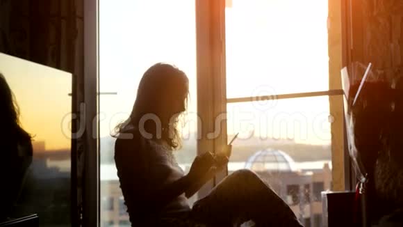 坐在窗户上的漂亮年轻女子在夕阳和城市的背景下使用智能手机3840x2160视频的预览图