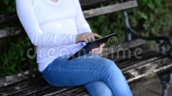 女人拿着数字平板电脑在公园的一张木凳上轻敲视频的预览图