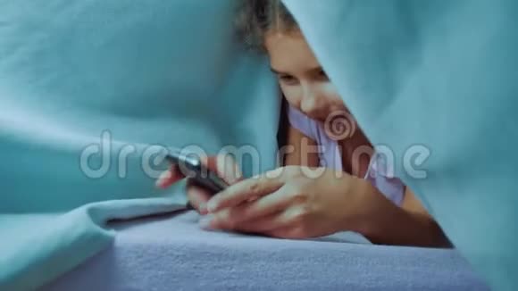 带智能手机的毯子下可爱女孩的肖像在社交媒体下玩网络生活方式游戏的小女孩视频的预览图
