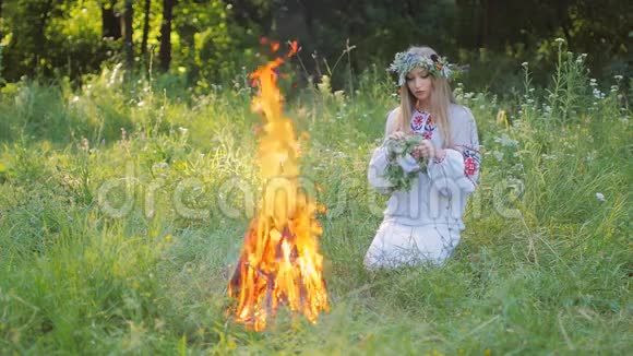 斯拉夫女人穿着刺绣的衬衫图案编织着一个野花的花环坐在火旁视频的预览图