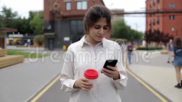 迷人的年轻女孩带着咖啡和智能手机在夏天的街道上散步视频的预览图