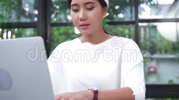 美丽的年轻微笑的亚洲女人在家客厅里一边在笔记本电脑上工作一边喜欢使用智能手机视频的预览图