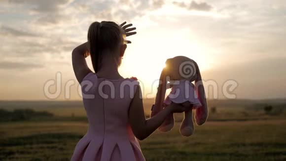 小女孩穿着粉红色的节日礼服在日落时拿着大毛绒兔子玩具一个带玩具的孩子的剪影纺织视频的预览图