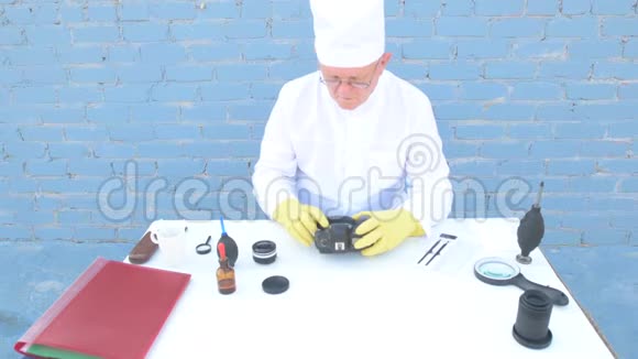 穿着白色长袍帽子医用口罩眼镜的老人用特殊手段清洁数码相机的基质女人视频的预览图