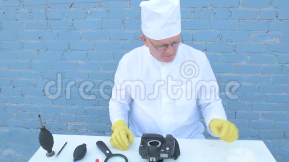 穿着白色长袍帽子医用口罩眼镜的老人用特殊手段清洁数码相机的基质女人视频的预览图