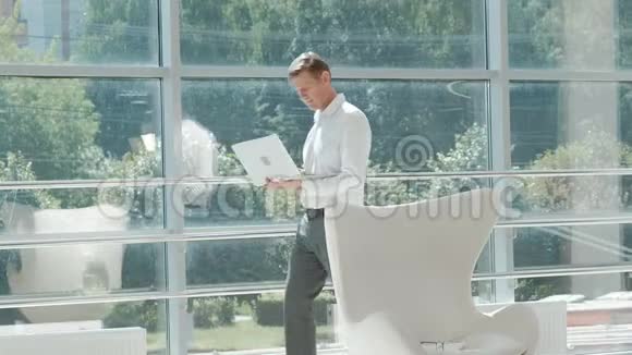 成熟的商人坐在玻璃办公室的白色椅子上用笔记本电脑工作视频的预览图