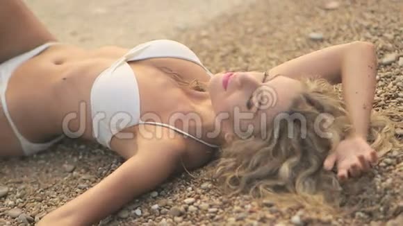 年轻漂亮的晒黑女孩穿着比基尼金色的头发躺在海边的日落视频的预览图