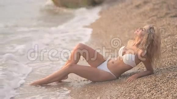 年轻漂亮的晒黑女孩穿着比基尼金色的头发躺在海边的日落视频的预览图