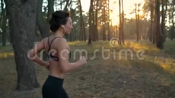 日落时分一个戴着耳机的女人在秋天的森林里奔跑慢动作视频的预览图