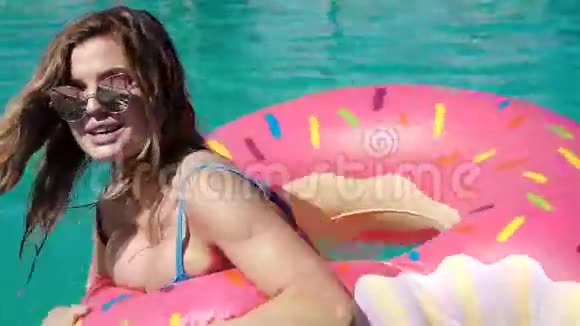 阳光明媚的夏日一位年轻貌美的女士戴着黑色比基尼的太阳镜躺在充气的粉红色甜甜圈里漂浮在游泳池里视频的预览图