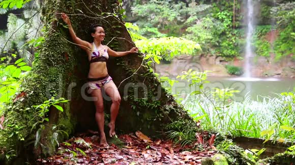 热带雨林背景瀑布的女孩斜倚着大树视频的预览图