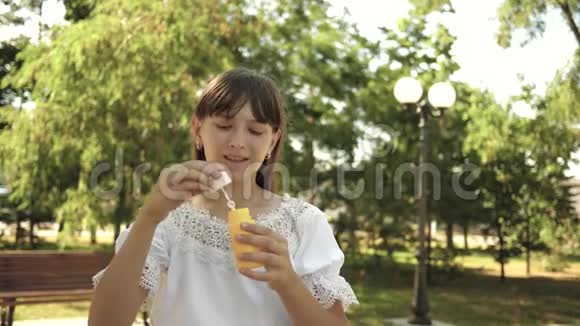 小女孩在公园里玩把泡泡吹进镜头慢动作漂亮的女孩吹肥皂视频的预览图