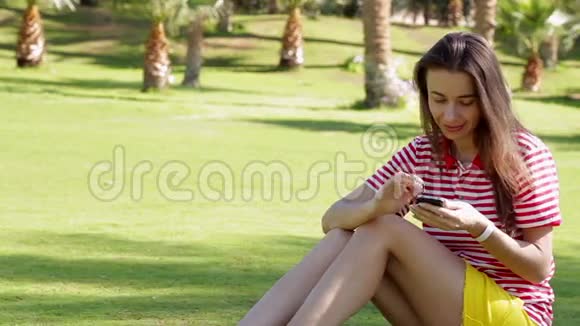 穿着黄色裙子坐在草地上打电话的年轻美女视频的预览图