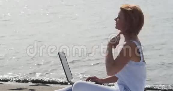 红头发的女孩坐在沙滩上的笔记本电脑上工作视频的预览图