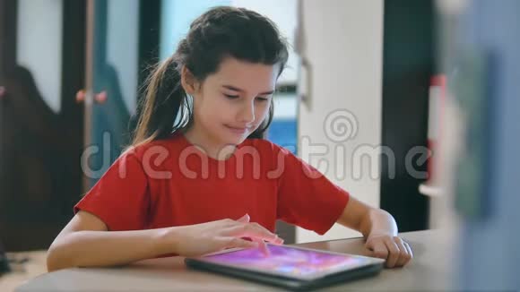 小女孩在玩数字平板电脑小女孩坐在桌子旁在网游社交媒体上玩数字平板电脑视频的预览图