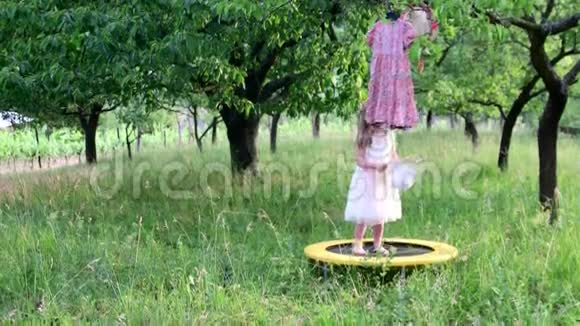 一个可爱的女孩在自然花园里跳舞小女孩在一个小蹦床上跳舞和跳跃小女孩穿白色衣服视频的预览图
