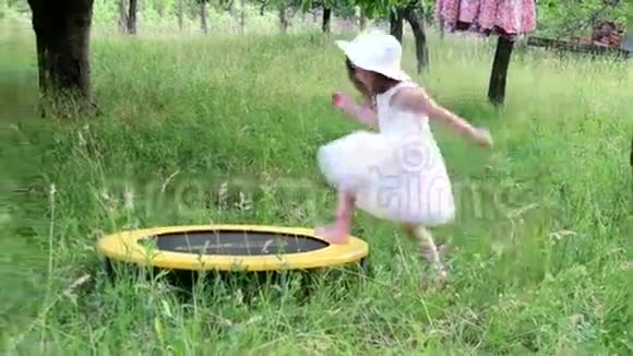一个可爱的女孩在自然花园里跳舞小女孩在一个小蹦床上跳舞和跳跃小女孩穿白色衣服视频的预览图