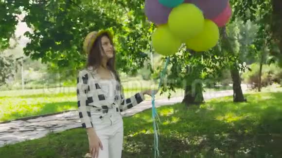 在阳光明媚的公园里美丽的女孩用五颜六色的气球在镜头前摆姿势的肖像视频的预览图