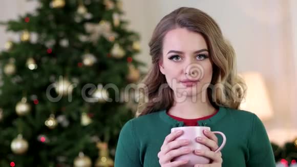 年轻漂亮的女人在圣诞树的背景上喝着杯子里的咖啡画着一幅美丽的肖像视频的预览图