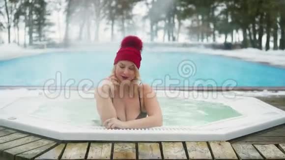 冬天在室外浴缸按摩浴缸里的红色小屋里的年轻金发女人视频的预览图
