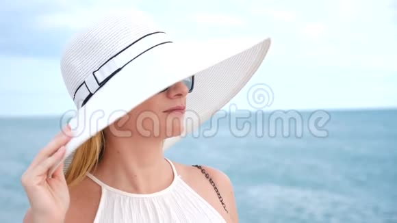 一位身穿白色宽边帽子和太阳镜的优雅女士欣赏海景4k慢镜头视频的预览图
