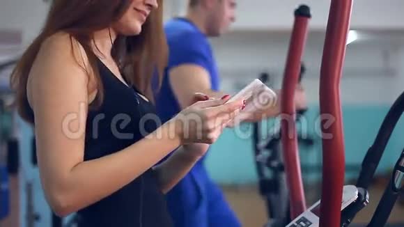 带着平板电脑的女孩运动员教练在模拟器上用椭球视频的预览图