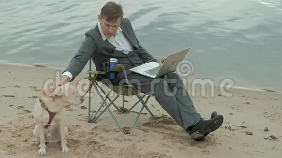 成熟的商人狗坐在河边放松穿西装用笔记本电脑的人视频的预览图