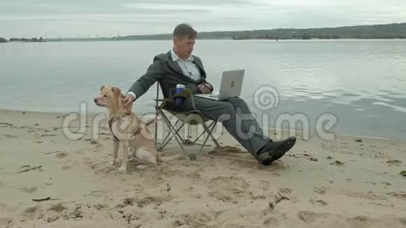 成熟的商人狗坐在河边放松穿西装用笔记本电脑的人视频的预览图