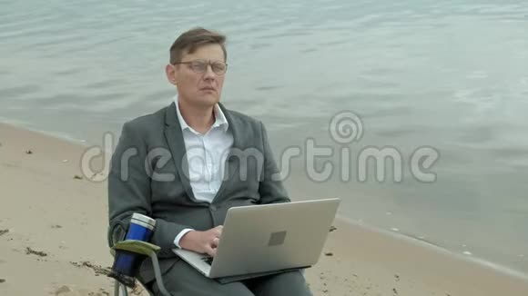 成熟的商人坐在河边放松穿西装用笔记本电脑的人视频的预览图