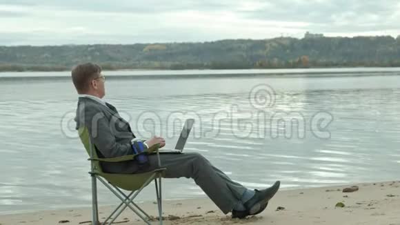 成熟的商人坐在河边放松穿西装用笔记本电脑的人视频的预览图