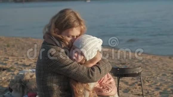 一个穿着外套的年轻女子和一个卷发的女孩一个母亲和一个女儿在河边的海滩上大海里野餐视频的预览图