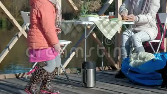 一个年轻的女人和一个穿着温暖衣服的小女孩母女在一座木桥上的河岸上野餐视频的预览图
