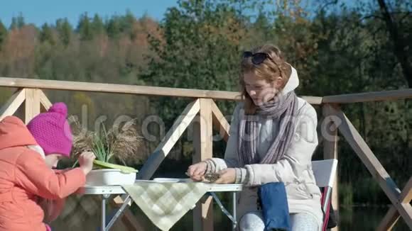 一个年轻的女人和一个穿着温暖衣服的小女孩母女在一座木桥上的河岸上野餐视频的预览图