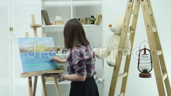 一位年轻女子站在画架后面的一张画布上在一个大气车间里画一幅画视频的预览图