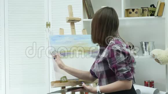 一位年轻女子站在画架后面的一张画布上在一个大气车间里画一幅画视频的预览图