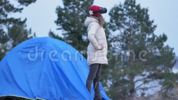 漂亮的年轻女游客戴着红帽子坐在旅游帐篷里享受着虚拟现实头盔视频的预览图