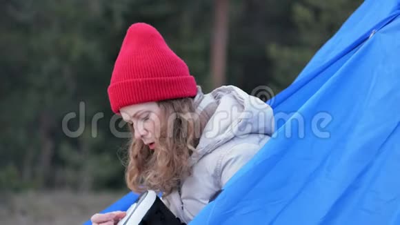 漂亮的年轻女游客戴着红帽子坐在旅游帐篷里享受着虚拟现实头盔视频的预览图