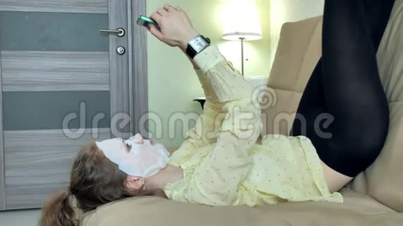 年轻女性在家里用智能手机点击沙发上的清洁面膜视频的预览图