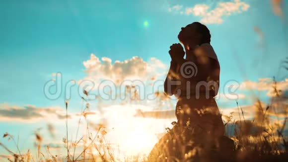 女孩在日落时双手交叉祈祷剪影女人跪着祈祷慢动作视频女孩折了起来视频的预览图