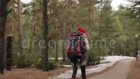 早春一个戴着红帽子的漂亮年轻女子背着一个大旅游背包穿过森林视频的预览图