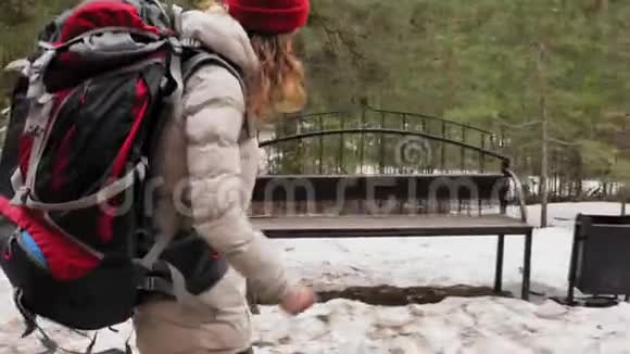 早春一个戴着红帽子的漂亮年轻女子背着一个大旅游背包穿过森林视频的预览图