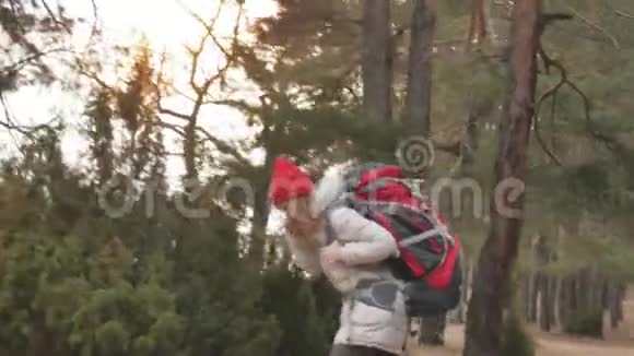 早春一个戴着红帽子的漂亮年轻女子背着一个大旅游背包穿过森林视频的预览图