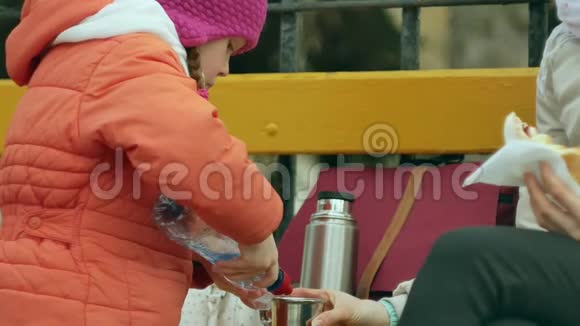 一个漂亮的女孩和她的妈妈坐在公园的长凳上吃着美味的甜点喝着热水瓶里的茶视频的预览图