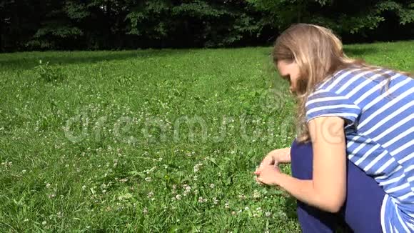 金发女人坐在草地上用三叶草植物的花编织头花环4K视频的预览图