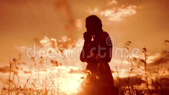 女孩在日落时双手交叉祈祷剪影女人跪着祈祷慢动作视频女孩折了起来视频的预览图