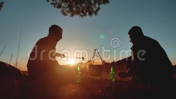 两名游客正坐在篝火晚会上在大自然中露营剪影阳光日落两个男人剪影视频的预览图