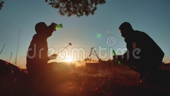 两名游客正坐在篝火晚会的露营生活方式中在大自然的剪影阳光下日落两个男人剪影视频的预览图