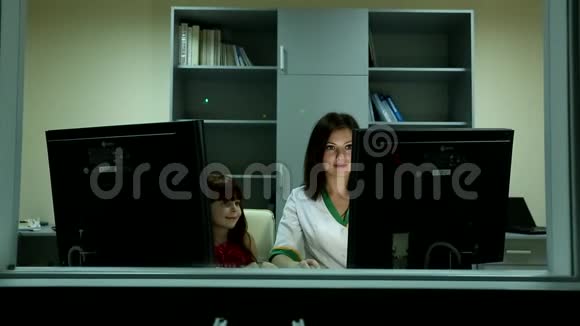 小可爱的女孩看着微笑年轻迷人的黑发女人医生背后两台电脑显示器商务办公室视频的预览图