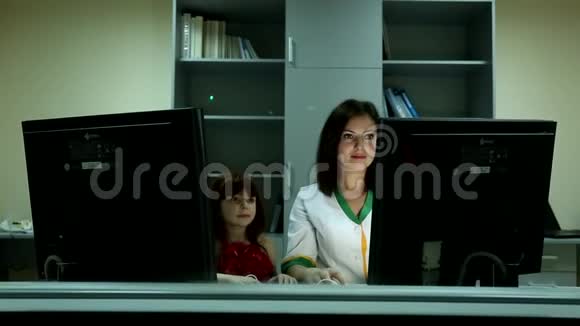 可爱的小女孩看着年轻漂亮的黑发女医生在商务办公室的两台电脑显示器后面微笑视频的预览图